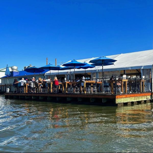 Outdoor seating on a dockside patio with blue umbrellas and people, adjacent to a body of water.