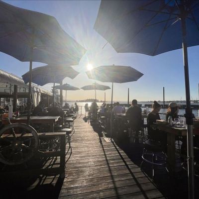 People dining outdoors under blue umbrellas with a sunset view over water.