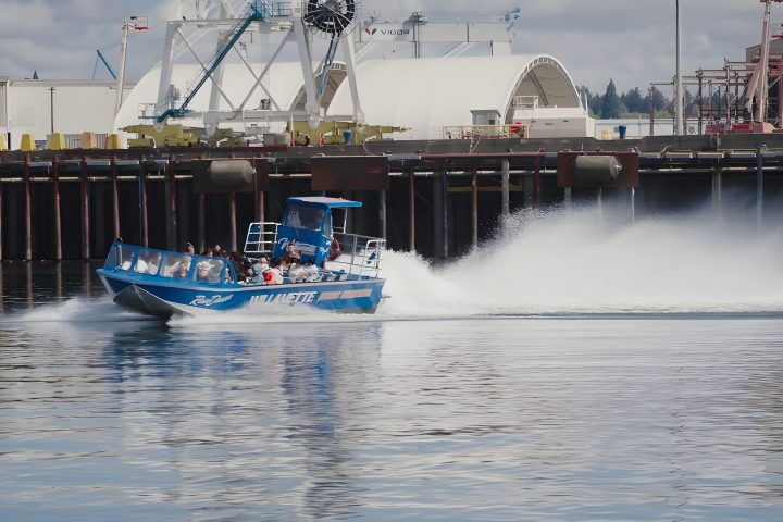 A blue speedboat with passengers speeding on a calm water surface near an industrial dock.