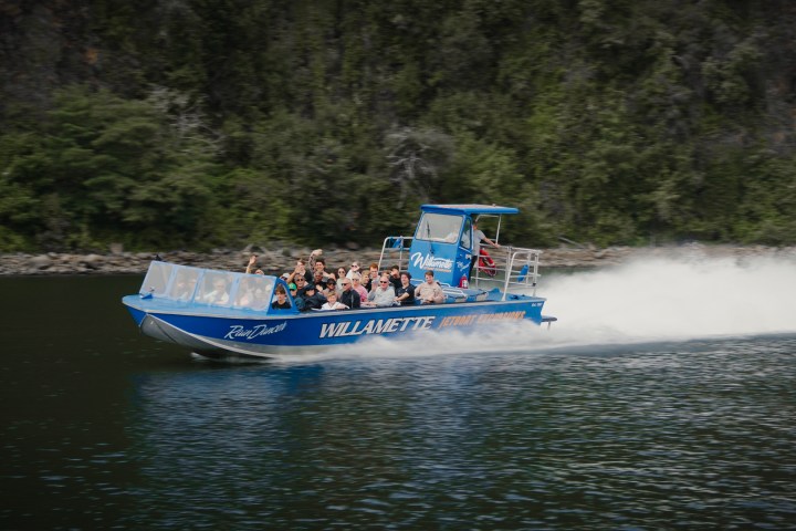 Blue jet boat with passengers speeding on a river near a forested shore.