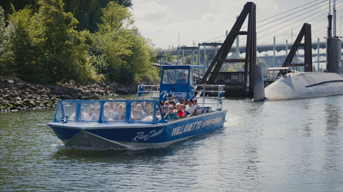 Tour boat with passengers on a river, near trees and a bridge.